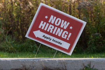 Outdoor lawn sign now hiring apply inside with a direction arrow, selective focus, blurred background. Employment and labor shortage, resigning, great resignation concept..