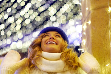 Smiling woman with garlands and holiday lights on festive Christmas or New Year fair. Lady wearing classic stylish winter knitted sweater and mittens