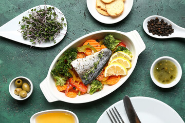Baking dish of tasty sea bass fish with vegetables and ingredients on green background