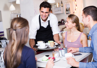 positive waiter bringing ordered dishes to friends in tearoom of a cozy confectioner's shop