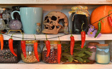 Wooden counters with witch ritual objects, cup, old bottles of herbs and potions, skull, hanging pepper and pumpkin in vintage kitchen.
