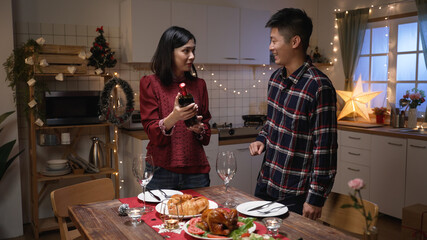 cheerful asian married couple standing by festive dinner table and discussing the Christmas wine they are going to have on the eve of holiday celebration