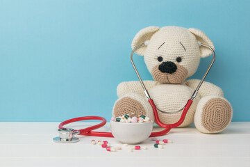 A red stethoscope on a white knitted bear and pills in a bowl.