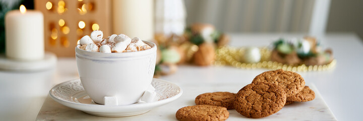 Long banner with cozy winter Christmas evening. Christmas holiday background with fir branch, cup of hot cocoa and marshmallow, candles on table. Kraft paper house. Winter hot drink, selective focus