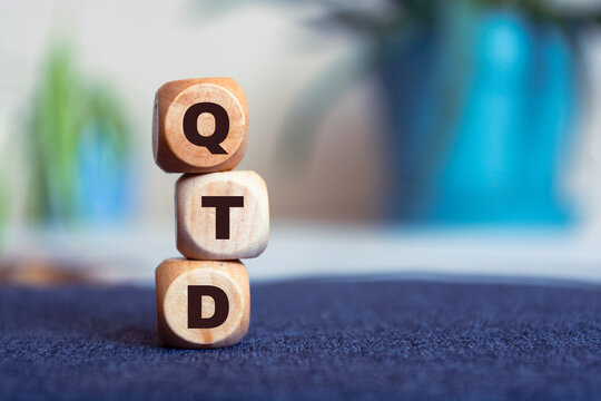 Motivational words: QTD in 3d wooden alphabet letters on a bright blur background with copy space, business concept. Front view concepts, flower in the background.