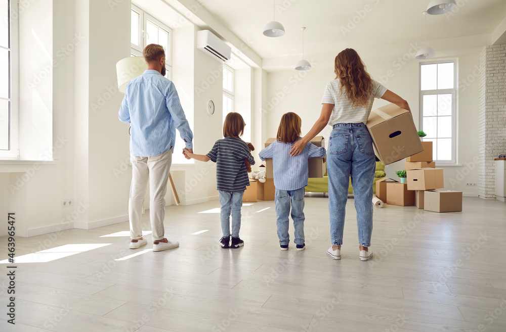 Wall mural Family standing in empty unfurnished spacious living room, rear view, from behind. Mom, dad and kids carrying and unpacking boxes together while moving into house or apartment. Buying new home concept