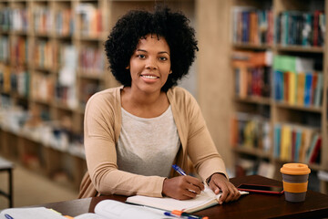 Happy mid adult black woman studying in library and looking at camera.