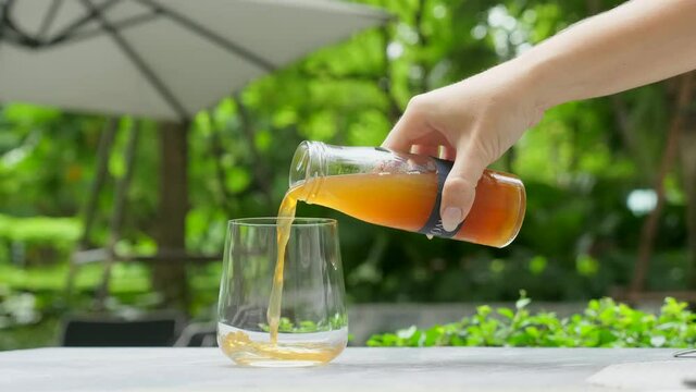 Woman hand pours kombucha drink, tea mushroom, tea fungus, manchurian mushroom, fermented tea from glass bottle to glass on marble table in outdoors restaurant or terrace cafe among green trees.