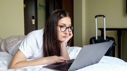 young woman in glasses looks at a laptop lying on the bed on stomach against the background of a...