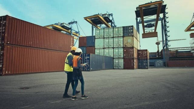 Multiethnic Female Industrial Engineer with Tablet and Black African American Male Supervisor in Hard Hats Walk in Container Terminal. VFX Double Girder Gantry Cranes Work in the Background.