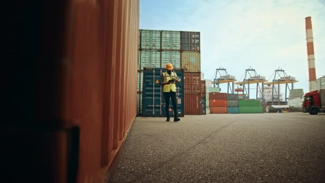 Black African American Male Industrial Supervisor Talking on Two-Way Radio and Holding a Tablet Computer while Walking in Container Terminal. VFX Double Girder Gantry Cranes Work in the Background.