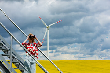 Woman in red and white striped jacket watching in binoculars with wind turbine on background in rapeseed field