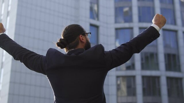 Cheerful and successful man in elegant suit with bunch on head rising hands up in winner gesture, looking at big corporate building, creating and developing his business empire, earning first billion