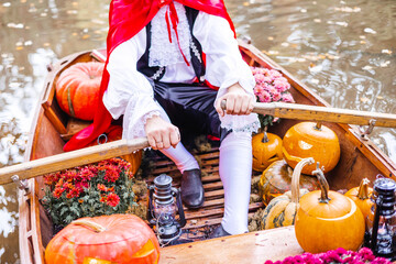 young man dressed as gondolier in gondola boat decorated with pumpkins on pond in autumn park celebrates Halloween and having fun, concept of Halloween carnival or costume party - obrazy, fototapety, plakaty
