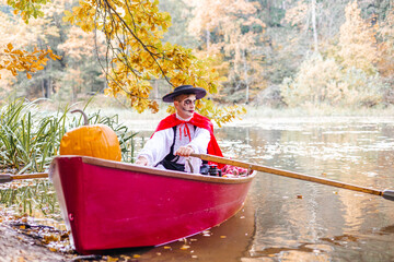 young man dressed as gondolier in gondola boat decorated with pumpkins on pond in autumn park celebrates Halloween and having fun, concept of Halloween carnival or costume party