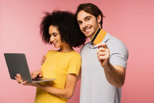 smiling hispanic woman using laptop near cheerful man holding credit card isolated on pink.