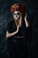 halloween redhead woman in makeup, witch skull costume calavera with sewn-on mouth, dried flowers in a smoky dark room, looking at the camera, touching the horns on her head