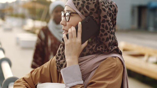 Chest-up of young Middle Eastern woman wearing hijab, talking on mobile phone, standing outdoors in city at daytime