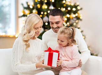 Fototapeta na wymiar family, winter holidays and people concept - happy mother, father and little daughter with gift box sitting on sofa at home over christmas tree background
