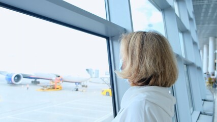 Female in a protective mask stands and looks out the window in the airport terminal awaiting the departure of a flight due to travel restrictions due to the coronavirus pandemic, a senior aged 50-55