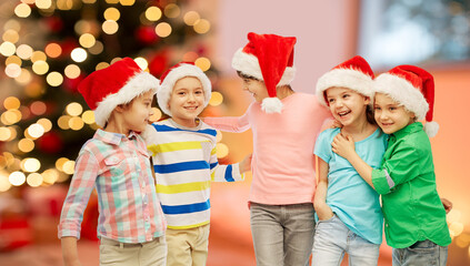 childhood, winter holidays and friendship concept - group of happy smiling little children in santa hats hugging over christmas tree lights background