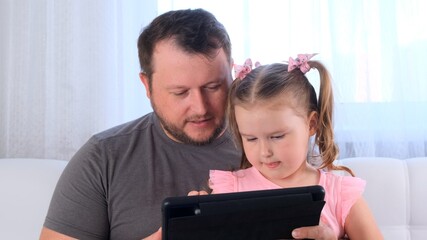 interested little girl 3 years old and her father study together on a tablet and watch a lesson online at home. Dad helps his daughter with her studies. Online school distance learning concept.