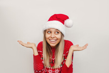 A young caucasian joyful smiling blonde woman in a red deer warm sweater and a Santa Claus hat spreads hands with doubt looking away isolated on a white background. Christmas and New Year's concept