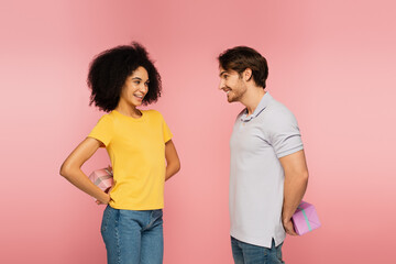 cheerful interracial couple looking at each other while holding presents behind back isolated on pink.