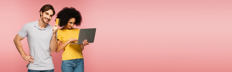 happy man showing credit card near hispanic girlfriend using laptop isolated on pink, banner.