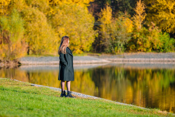 Naklejka na ściany i meble A woman with long hair walks in an autumn park by the pond. Autumn season.