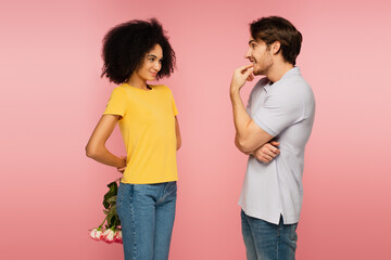 curious man looking at smiling latin girlfriend holding bouquet behind back isolated on pink.