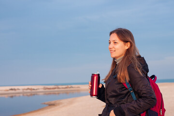 Happy young woman in black raincoat with backpack on empty beach autumn sea. Smiling millennial girl with long hair walking hiking drinking tea in thermo can bottle alone.Lifestyle real people outdoor