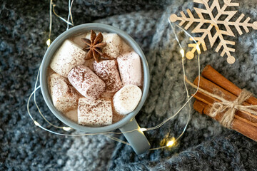 Winter still life from fir branches, scarf,grey mug of cocoa, coffee or hot chocolate with marshmallow on warm plaid with garland. Concept of cozy christmas home environment. Top view