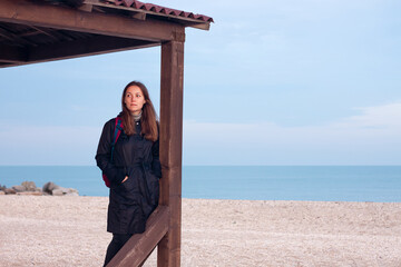 Happy pretty young woman in black raincoat with backpack on empty beach autumn sea. Smiling millennial girl with long hair walking hiking resting alone. Lifestyle real people female portrait outdoor