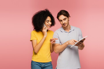 hispanic woman talking on smartphone and pointing at digital tablet together with boyfriend isolated on pink.