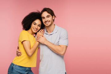 young and happy multiethnic couple embracing while smiling at camera isolated on pink.