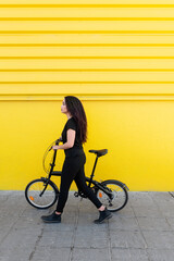 Stylish young black woman with long hair carrying her folding Bicycle In Front Of A Yellow Wall