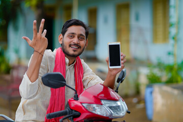 technology concept : Young indian farmer sitting on bike and showing smartphone.