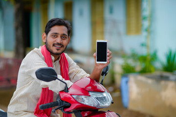 technology concept : Young indian farmer sitting on bike and showing smartphone.