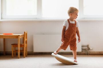Cute toddler child boy swinging on a balancing board in a light room, physical and sensory...