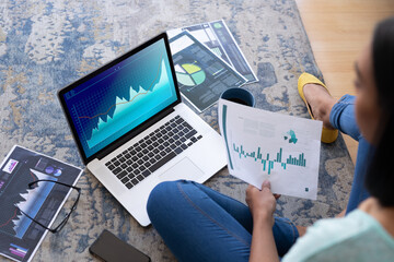 Biracial woman sitting on floor, using computer with statistical data on screen