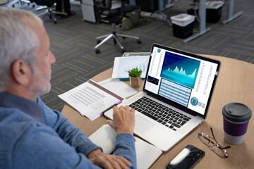 Caucasian senior businessman sitting at desk, using laptop with statistical data on screen