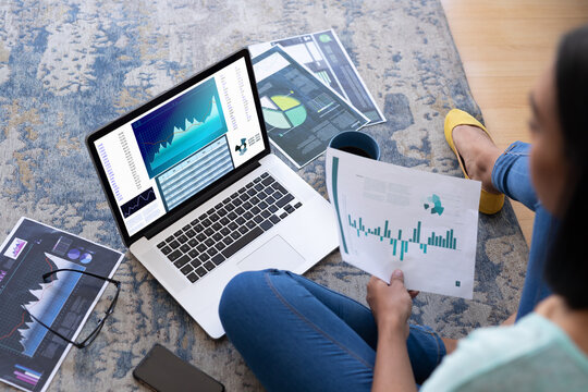 Biracial businesswoman sitting on floor using laptop with statistical data on screen