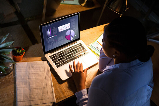 Biracial businesswoman sitting at desk using laptop with statistical data on screen
