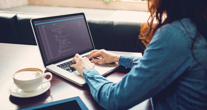 Caucasian female programmer sitting at desk with coffee, using laptop with coding on screen