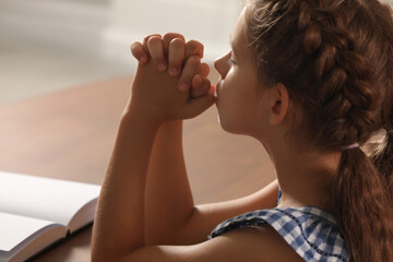 Cute little girl praying over Bible at table indoors, closeup