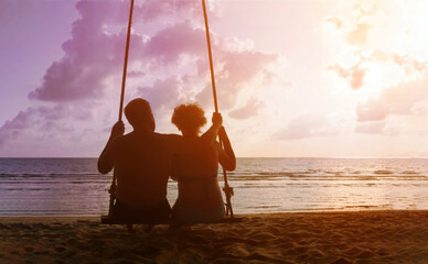 Adult retired couple sitting on rope swing and looking on sunset sea beach. Man embrace his wife. Mature family relax on vacation.
