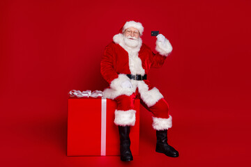 Photo of handsome positive retired man wear santa claus costume spectacles smiling buying credit card present isolated red color background