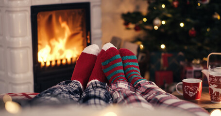 Couple feet in cozy christmas woolen socks near fireplace with decorated xmas tree and tee cup in background - obrazy, fototapety, plakaty