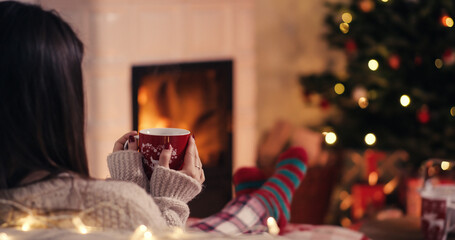 Over the shoulder young woman relaxing near fireplace with cup of tee in cozy christmas woolen socks with decorated xmas tree in background